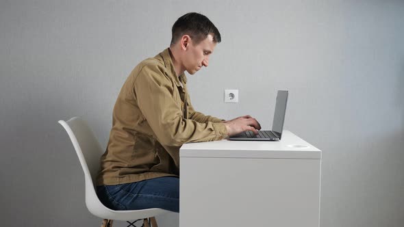 Young Man Fidgets on Chair Working Via Laptop at Table