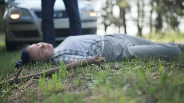 Blurred Knocked Down Pedestrian Lying on Green Grass with Legs Walking at Background