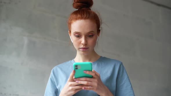 Attractive Redhead Young Woman Wearing Wireless Earphones Is Listening Music Typing Message on Cell.