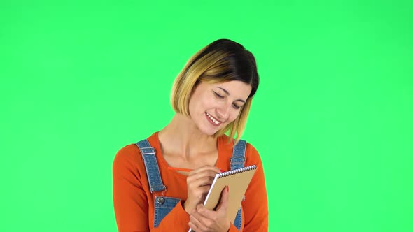 Girl Stands and Thinks, Then Happy Writes with Pencil in Notebook. Green Screen