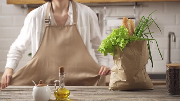 Woman Chef Puts On An Apron For Camera. The First Step In Food Preparation. There Is A Paper Bag