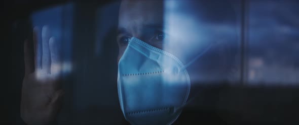 Man standing with his hand on a window looking out and taking off his medical mask