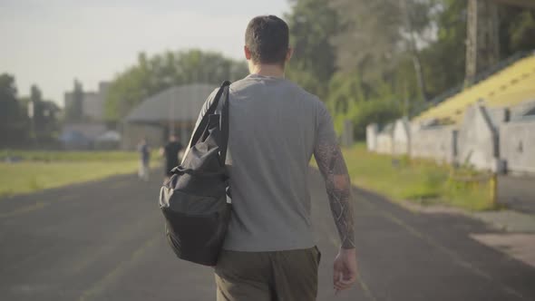 Back View of Confident Caucasian Man with Sport Bag Walking Along Stadium in Summer Morning. Camera