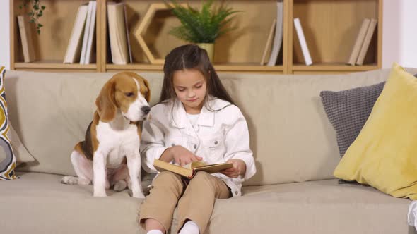 Little Girl Sitting with Dog on Sofa and Reading Book