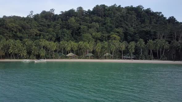 Aerial view above turquoise waters towards green tropical island with palm trees and huts in the Phi