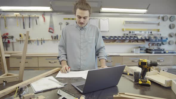 Caucasian Carpenter Working with Laptop and Technical Drawings at Workshop