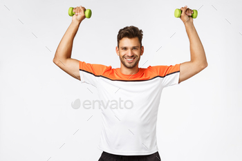  muscular guy with bristle in sportswear lift hands up, holding dumbbells smiling carefree, proud with body shape, got fit for beach season.