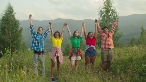 Happy Multiethnic Tourists Standing with Arms Raised on Mountain Top at Sunset