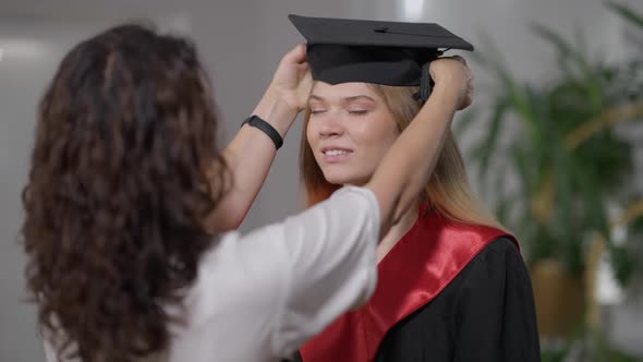 Portrait of Smiling Slim Beautiful Young Woman Looking at Mother Putting on Graduation Hat