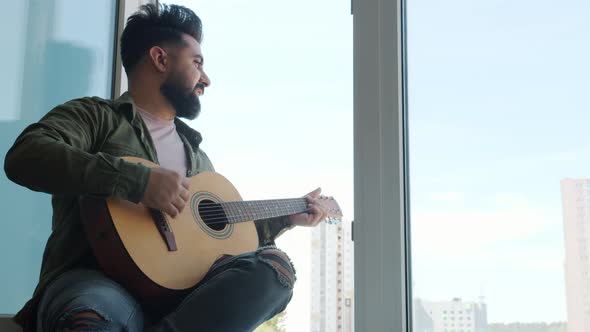 Slow Motion of Young Middle Eastern Man Playing the Guitar Singing Song Sitting on Window Sill at