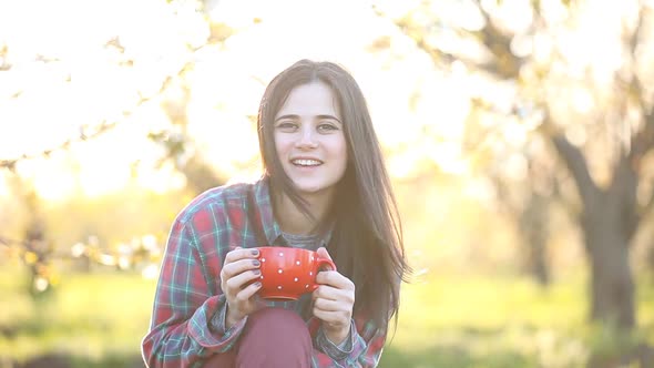 Brunet girl in shirt with a cup next to an apple tree garden.