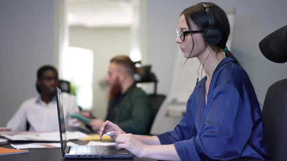 Side View Young Concentrated Woman in Headphones Talking Typing on Laptop Keyboard with Blurred Men