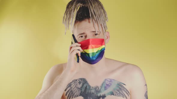 Naked young man with tattoo on chest in LGBT mask talking on mobile phone.