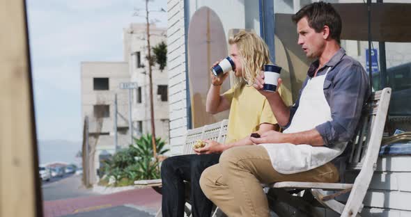 Two Caucasian male surfboard makers sitting on a bench and drinking takeaway coffee