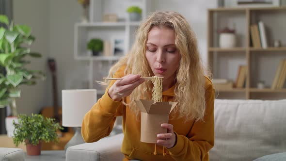 Young Woman Eating Noodles From a Box with Chopsticks at Home on a Couch