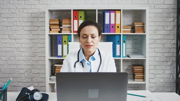 Female Doctor is Sitting at Table in Office of Modern Clinic and Consulting Her Patient By Laptop