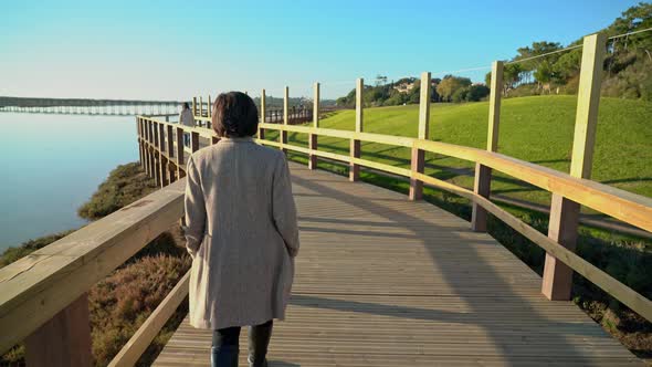 Two Females Walking on a Boardwalk Near a River Shore with People Ahead