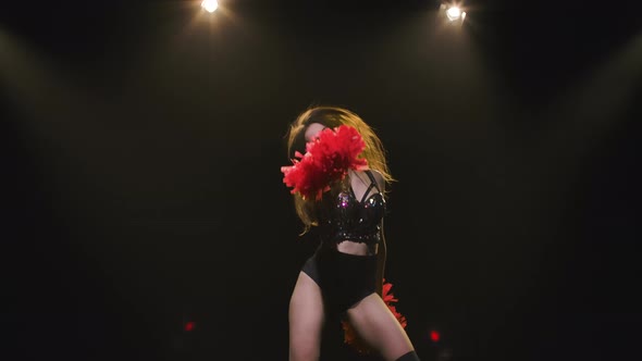 A Cute Cheerleader with Pompoms is Dancing on a Black Studio Background with Lights and Smoke