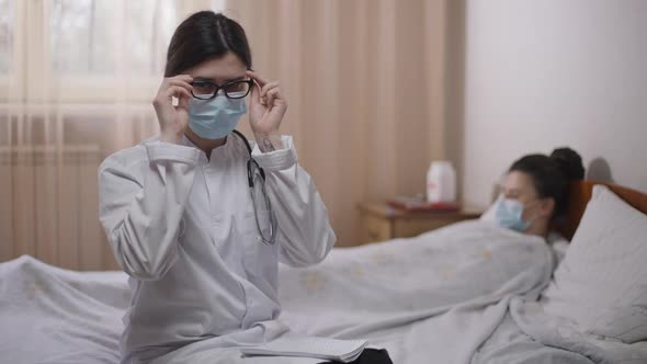 Portrait of Female Caucasian Doctor in Covid Face Mask Putting on Eyeglasses Looking at Camera