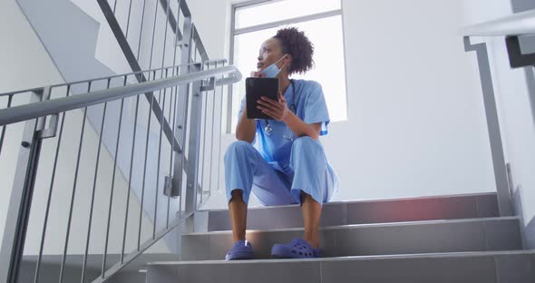 Mixed race female doctor wearing face mask sitting on stairs in hospital using tablet