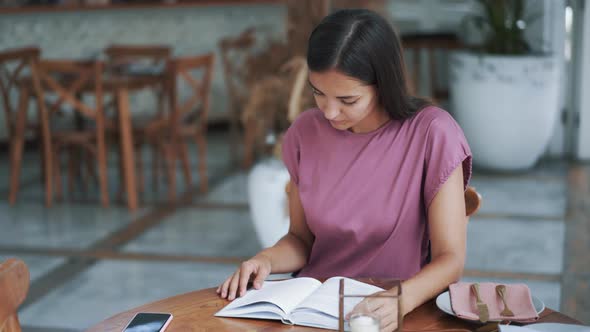 Portrait of Beautiful Woman Sitting at Table in Cafe and Reading Book