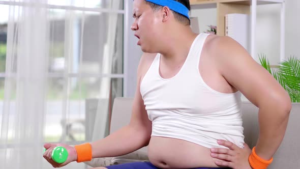 Fat Asian guy in sportswear lifting one side dumbbell with difficultly in living room