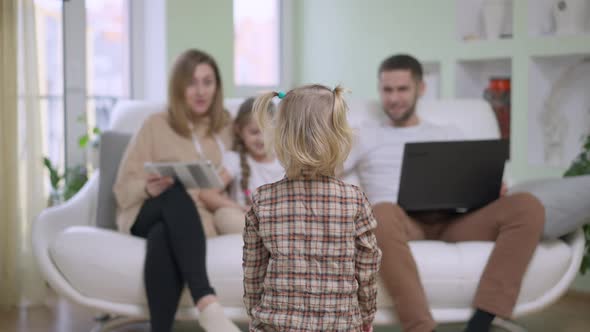 Back View of Blond Caucasian Baby Girl Walking to Family Sitting on Couch at Home