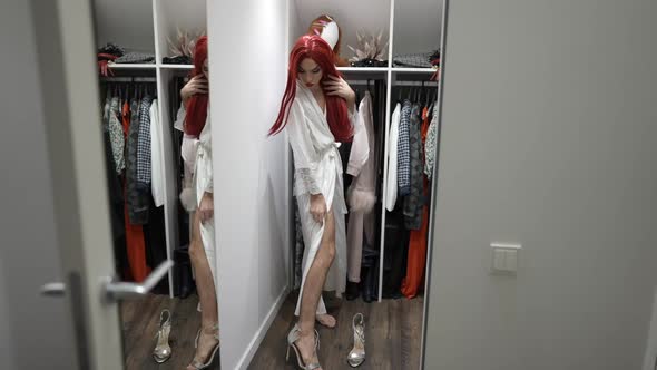 Wide Shot Thoughtful Transgender Woman in White Silk Bathrobe and Red Wig Trying on Luxurious