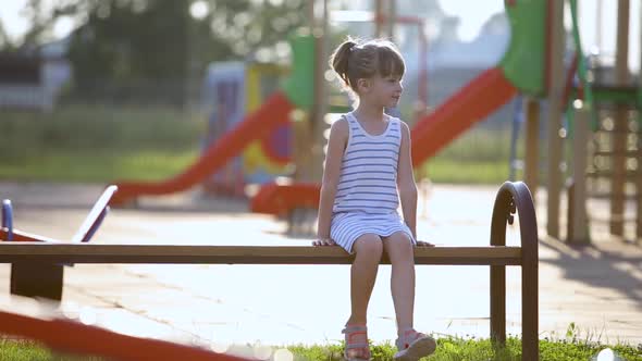 Cute Child Girl Waiting for Her Mother Sitting on a Bench on Summer Playground in Kindergarten Yard