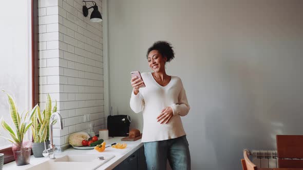 Happy African pregnant woman looking at phone and dancing