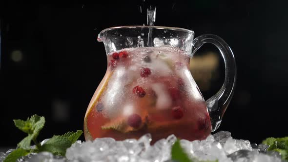 Pouring Pure Sparkling Cold Water Into Transparent Glass Jar with Lime Ice Cubes and Berries