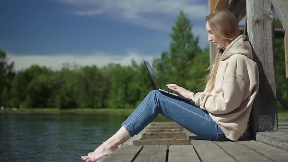 Beautiful Young Girl is Using Laptop and Typing While Sitting on Pier By Lake on Summer Day Spbd