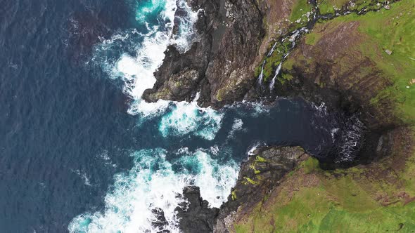 Aerial View of Waves Break on Rocks of Faroe Islands Cliffs in a Blue Ocean