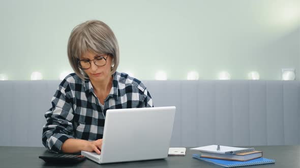 An Elderly Tired Woman with Glasses is Sitting at a Laptop and with Difficulty Overcoming Drowsiness