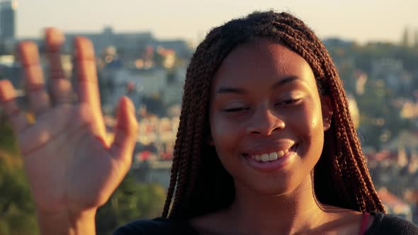 Young Beautiful Black Woman Waves with Hand To Camera - City in the Background