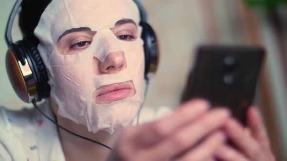 Portrait, Young Woman in White Cosmetic Fabric Mask, with Headphones on Head, Holds Smartphone