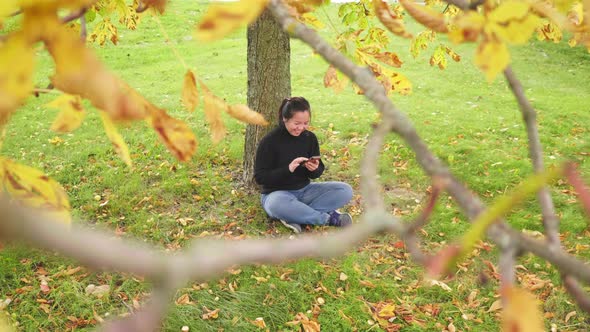 Happy Asian woman sitting and slide the screen on smartphone under tree at the park in Autumn