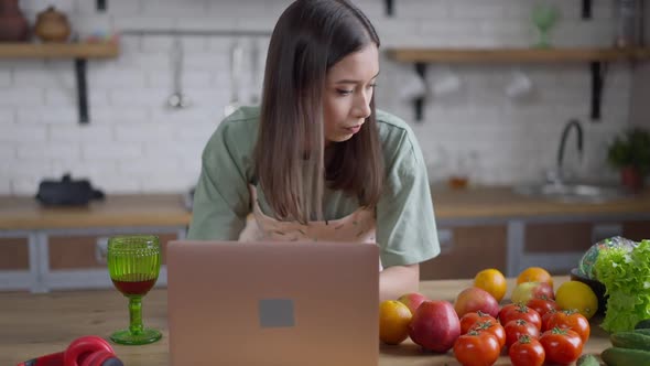 Concentrated Young Asian Woman Typing on Laptop Keyboard Checking Ingredients for Homemade Dish and