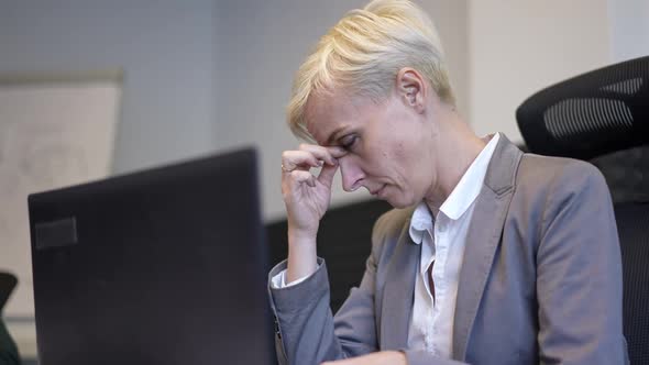 Overburdened Caucasian Woman in Formal Suit Rubbing Eyes Typing on Laptop Keyboard Sitting at Table