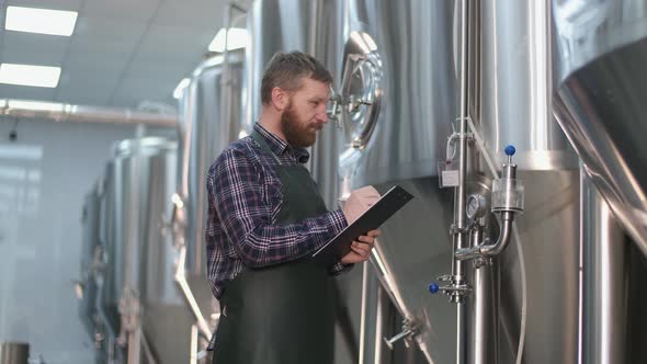 A Male Brewer in an Apron with a Beard Stands Near the Beer Tanks and Writes Down the Readings