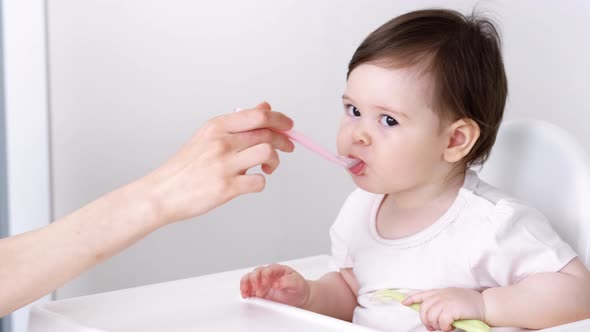 Baby Girl Eating Blend Mashed Food Sitting on High Chair Mother Feeding Child Hand with Spoon for