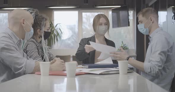 Confident Young Woman in Covid Face Mask Passing Documents To Coworkers at Meeting