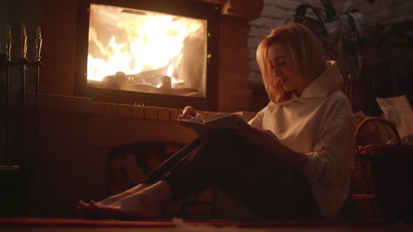 A Young Blonde Woman is Reading a Book Near Fireplace