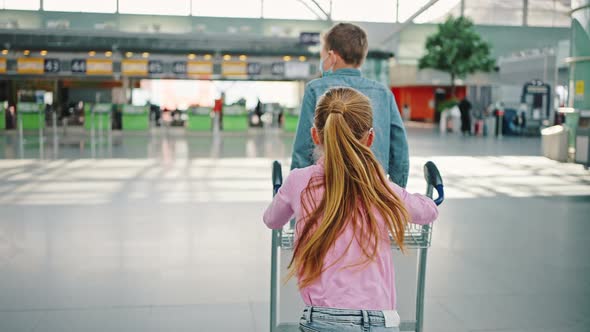 Follow Shot of Two Kids Fooling Around at Airport Hall Running with Luggage Cart Back View