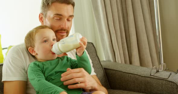 Boy in fathers lap drinking milk on sofa 