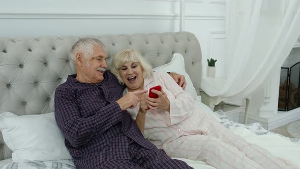 Senior Elderly Couple Wearing Pyjamas Lying on Bed Looking on Mobile Phone Making Online Shopping
