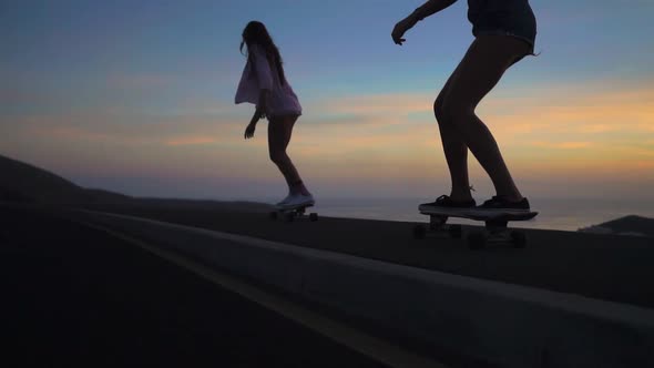 Two Girls on Skateboards Ride on the Road Against the Rock and Sky During Sunset