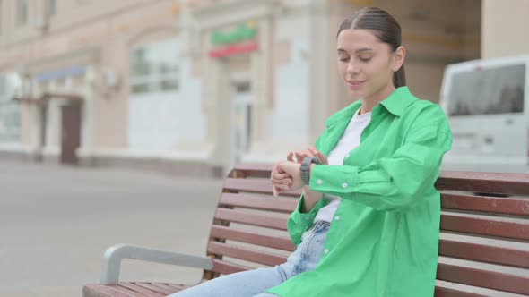 Hispanic Woman Using Smart Watch While Sitting on Bench Outdoor