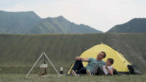 Happy Father and Son Relax Together and Rejoice in Front of the Tent Against the Backdrop of the