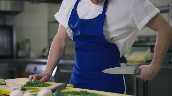 Unrecognizable Male Cook Cleaning Kitchen Knife Rubbing Utensil on Apron and Cutting Greenery